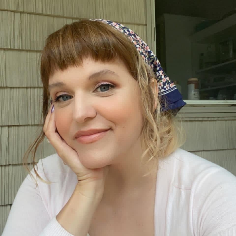 Libby looks at the camera with a smile on her face, resting her face on her hand. Her nails are metallic purple and her hair is pulled back in a bandana.